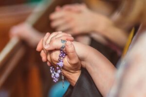 Close-up of hands clasped in prayer holding a purple rosary in a church setting.